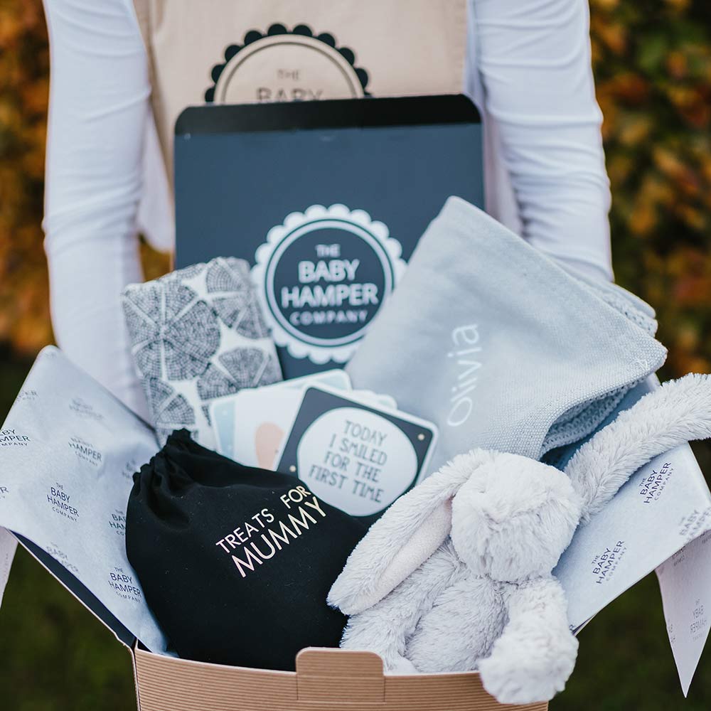 Baby gift hamper with items including a blanket, card, and small plush toy, branded by The Baby Hamper Company.