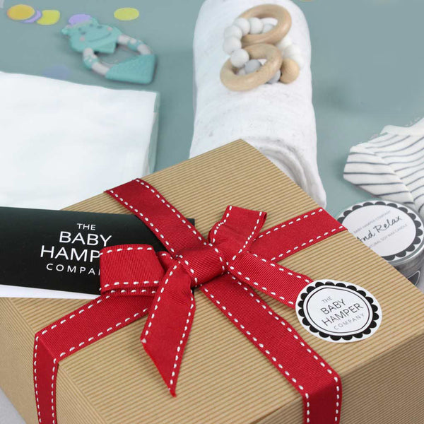 Gift box with a red ribbon and 'The Baby Hamper Company' label, surrounded by baby items on a light blue background.